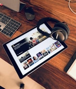A wooden table holds a tablet displaying a streaming service interface with various movie and TV show thumbnails. Nearby, a pair of Bose headphones rests on the table along with a keyboard and a mouse.