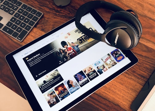A wooden table holds a tablet displaying a streaming service interface with various movie and TV show thumbnails. Nearby, a pair of Bose headphones rests on the table along with a keyboard and a mouse.