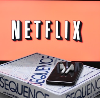 A screen displaying the Netflix logo in bold, black and white lettering against a vibrant red background. In front of the screen, there is a closed box for the board game 'Sequence', with a smartphone and wireless earbuds resting on top.
