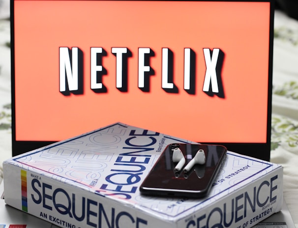 A screen displaying the Netflix logo in bold, black and white lettering against a vibrant red background. In front of the screen, there is a closed box for the board game 'Sequence', with a smartphone and wireless earbuds resting on top.