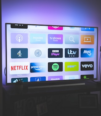 A flat-screen television is displayed in a dimly lit room, showcasing a variety of streaming service apps on its screen, such as Netflix, Prime Video, and Spotify. The TV is positioned on a black stand beside a tall floor lamp, casting a soft ambient light on the surrounding area.