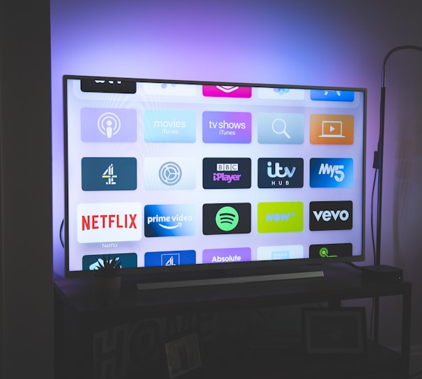 A flat-screen television is displayed in a dimly lit room, showcasing a variety of streaming service apps on its screen, such as Netflix, Prime Video, and Spotify. The TV is positioned on a black stand beside a tall floor lamp, casting a soft ambient light on the surrounding area.