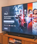 A television displaying a Disney+ promotional screen featuring various characters from popular franchises. Below the screen is a wooden cabinet with a few books and a small plant. The wall is light-colored, creating a cozy and inviting atmosphere.