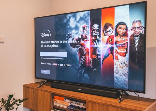 A television displaying a Disney+ promotional screen featuring various characters from popular franchises. Below the screen is a wooden cabinet with a few books and a small plant. The wall is light-colored, creating a cozy and inviting atmosphere.
