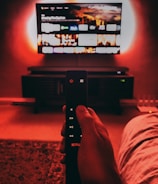 A hand holds a remote with visible buttons, pointed towards a wall-mounted TV displaying a streaming platform interface. The room is dimly lit, with a warm, red ambient light highlighting the TV screen and surroundings.