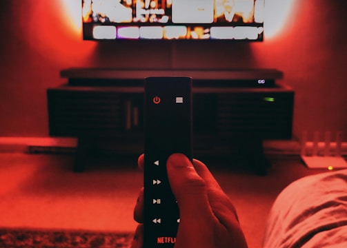 A hand holds a remote with visible buttons, pointed towards a wall-mounted TV displaying a streaming platform interface. The room is dimly lit, with a warm, red ambient light highlighting the TV screen and surroundings.