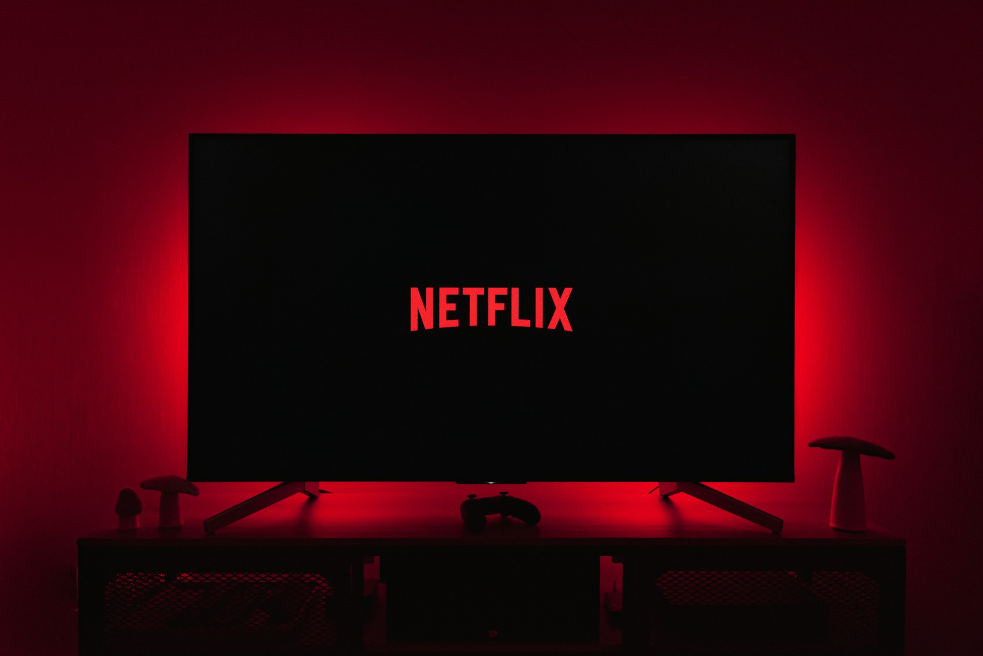 A dark room featuring a television screen displaying the word 'Netflix' in red against a black background. The screen is centered on a console table, with a video game controller placed in front. The room is dimly lit by red ambient lighting, creating a cozy atmosphere.