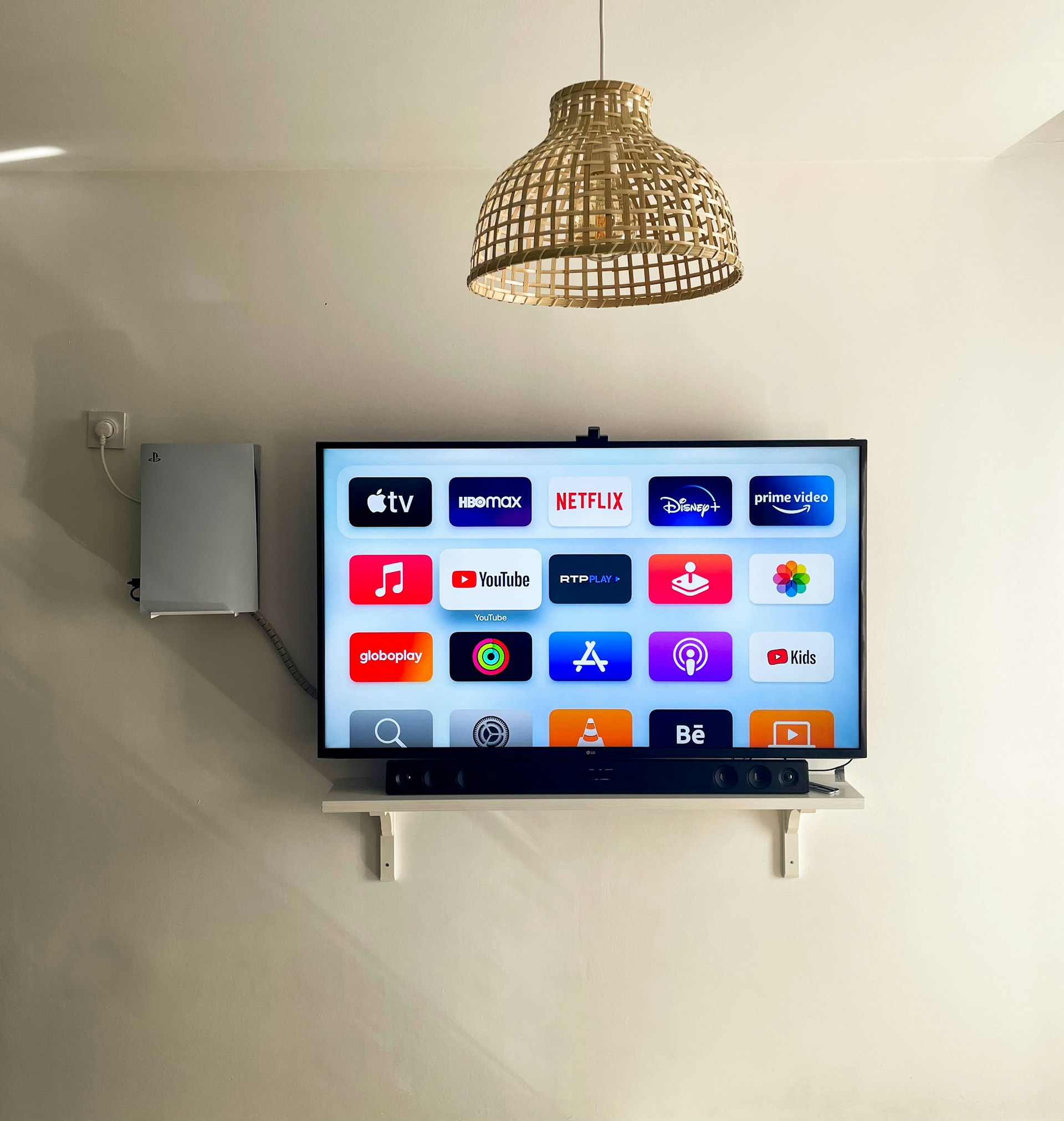 A modern living room features a wall-mounted flat-screen television displaying multiple streaming app icons, such as Apple TV, HBO Max, Netflix, Disney+, and Prime Video. Below the TV is a soundbar sitting on a small white shelf. A wicker pendant light hangs from the ceiling above, and the wall is painted a neutral color.