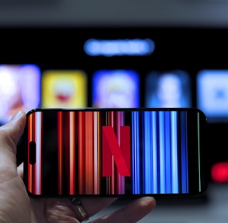 A hand holding a smartphone displaying the Netflix logo on its screen with a background of colorful vertical lines. In the background, a television screen shows blurry icons or profiles with various colors.
