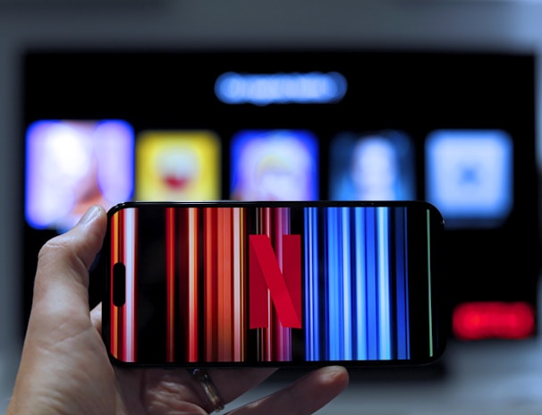 A hand holding a smartphone displaying the Netflix logo on its screen with a background of colorful vertical lines. In the background, a television screen shows blurry icons or profiles with various colors.