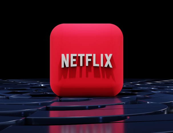 A red, cube-shaped icon with the word 'NETFLIX' prominently displayed in white, bold letters. The cube is set against a dark, glossy, circular patterned background, reflecting a sleek, futuristic design.