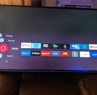 A television screen displays a smart TV interface with multiple app icons arranged in rows. Options such as Settings, Sources, Home, Search, and TV are listed on the left side. Some recognizable app icons include Netflix, Prime Video, BBC iPlayer, ITV Hub, All 4, My5, YouTube, and TikTok. The time and date are shown in the top right corner.