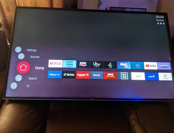 A television screen displays a smart TV interface with multiple app icons arranged in rows. Options such as Settings, Sources, Home, Search, and TV are listed on the left side. Some recognizable app icons include Netflix, Prime Video, BBC iPlayer, ITV Hub, All 4, My5, YouTube, and TikTok. The time and date are shown in the top right corner.