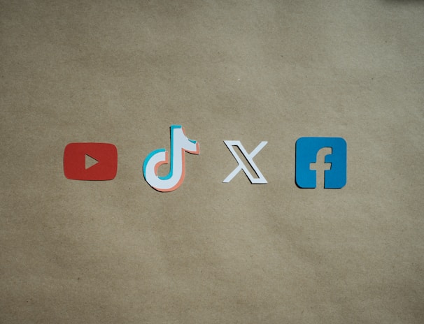 Four social media platform logos are arranged on a brown background. From left to right, the logos are for YouTube in red, TikTok in a combination of red, blue, and white, X in white, and Facebook in blue.